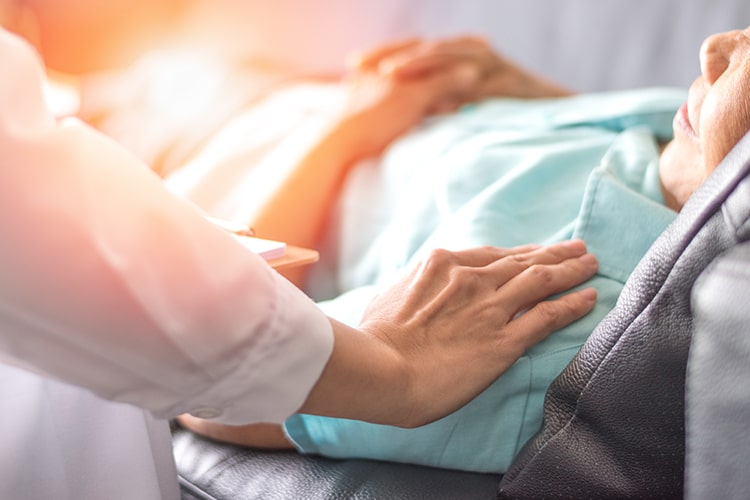 Elderly woman laying in hospital bed while practitioner consoles her by putting their hand on her shoulder