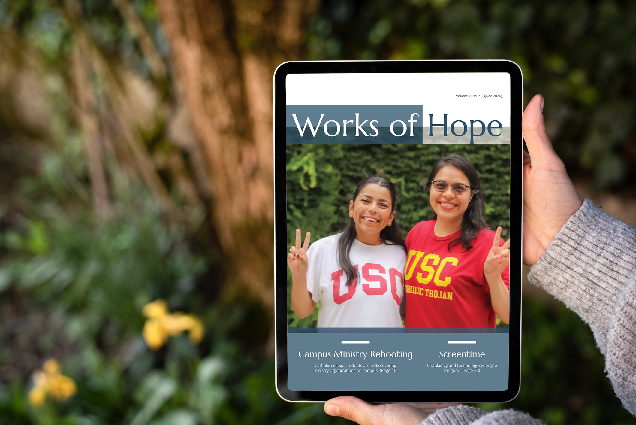 A tablet displaying the cover of Works of Hope magazine, Volume 2, Issue 2 (June 2026). The cover features two smiling young women wearing USC (University of Southern California) shirts — one white, one red — both making peace signs. The masthead reads "Works of Hope" in large text against a dark teal banner. Below the photo, two article teasers appear: "Campus Ministry Rebooting — Catholic college students are rediscovering ministry organizations on campus (Page 46)" and "Screentime — Chaplaincy and technology synergize for good (Page 36)." The tablet is held against a blurred outdoor background with green foliage and yellow flowers.